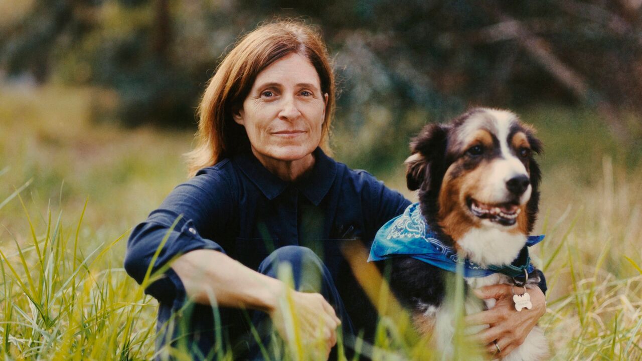 2025 Artist Fellowship grantee Jona Frank sits in a field of tall grass with her dog, wearing a blue bandana around its neck